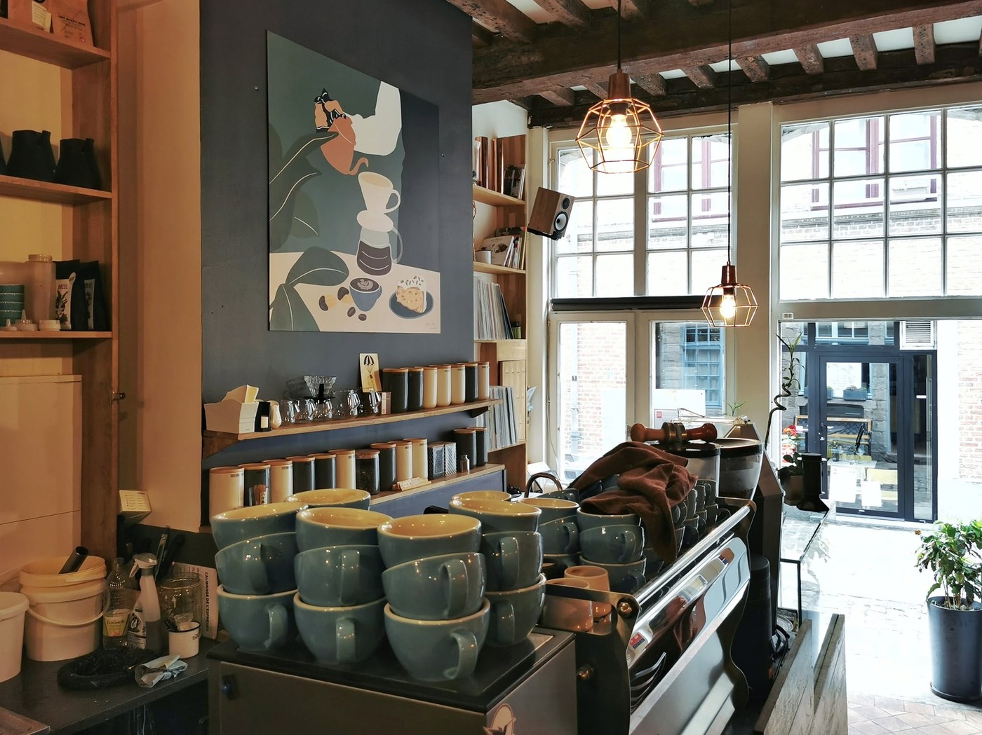 Stacked cups on the espresso machine beneath a shelf of coffee canisters and a pour over illustration, exposed wood beams and pendant lights overhead