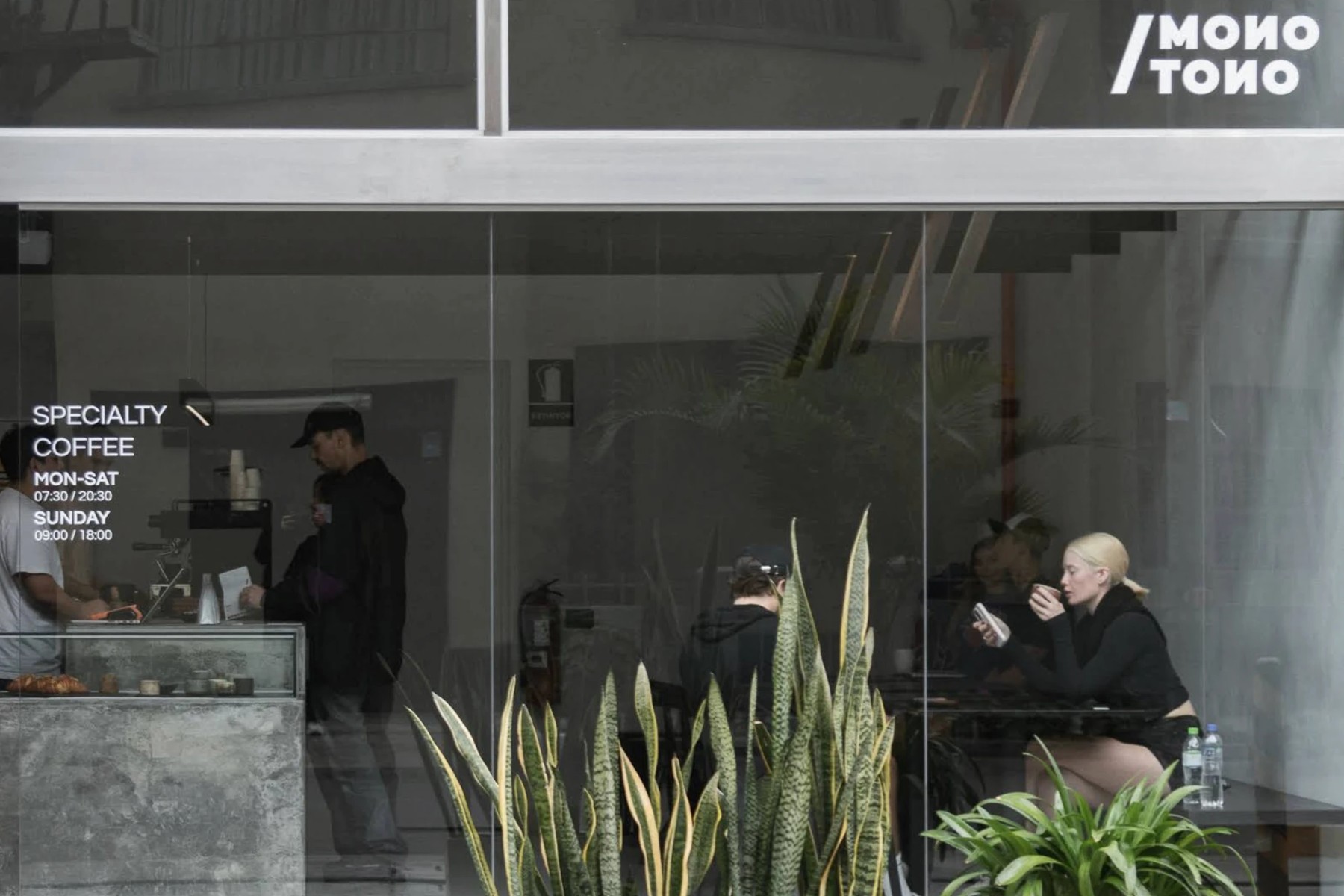 The glass front of Monotono Coffee with the white Monotono logo above, specialty coffee and opening hours on the window, a concrete pastry counter on the left and snake plants in front