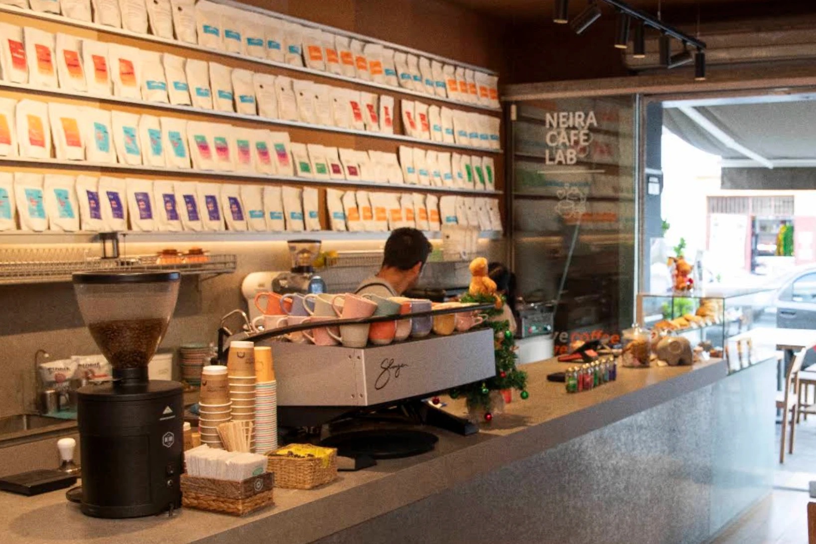 The counter at Neira Cafe Lab with a wall of white coffee bags in coloured labels, a silver espresso machine topped with pastel cups, a grinder and the Neira Cafe Lab lettering on the glass