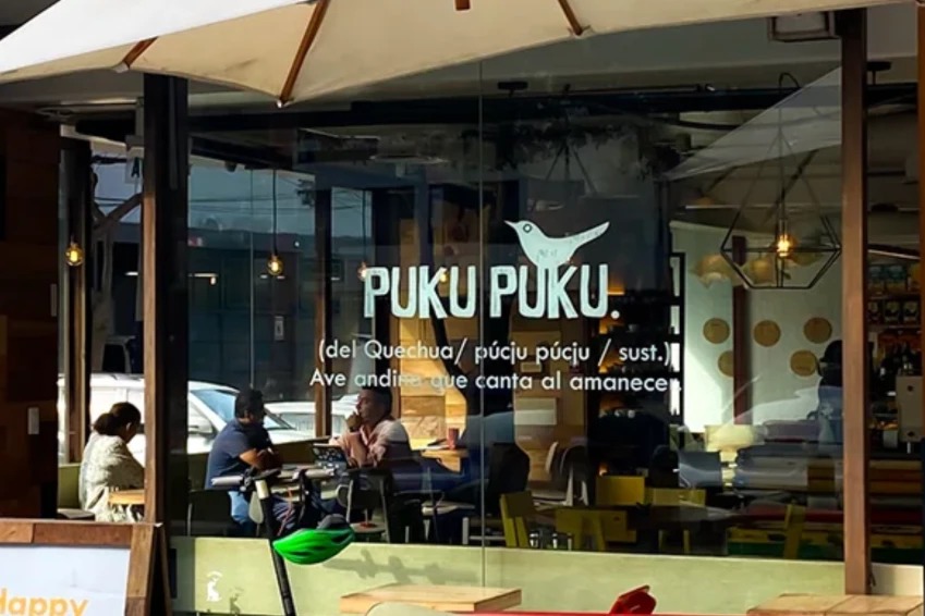 The glass front of Puku Puku with the white bird logo and Puku Puku name on the window, diners at tables inside and a cream parasol over the terrace