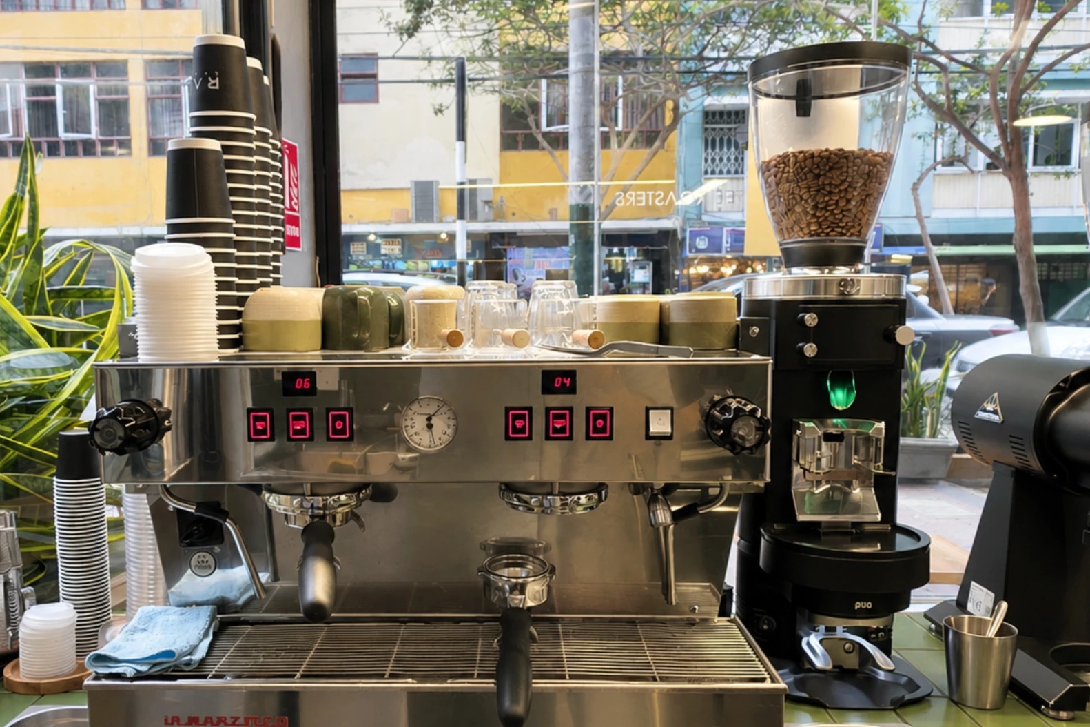 A chrome two group espresso machine topped with stacked cups, beside a grinder full of beans, in front of a street window with a snake plant and green tiled counter