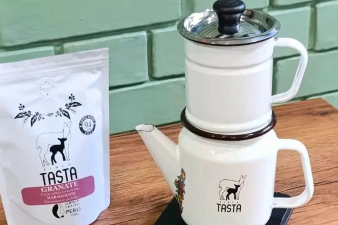 A bag of Finca Tasta Granate coffee beside a white enamel two part coffee brewer with the Finca Tasta deer logo, on a wooden table against a mint green brick wall