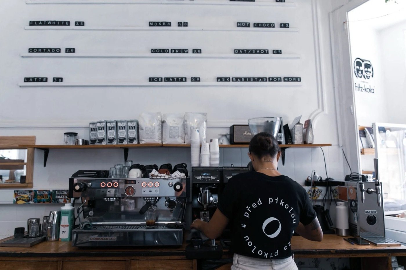 A barista pulling a shot behind the counter at Buna, letterboard menu and bagged coffee visible on the shelf