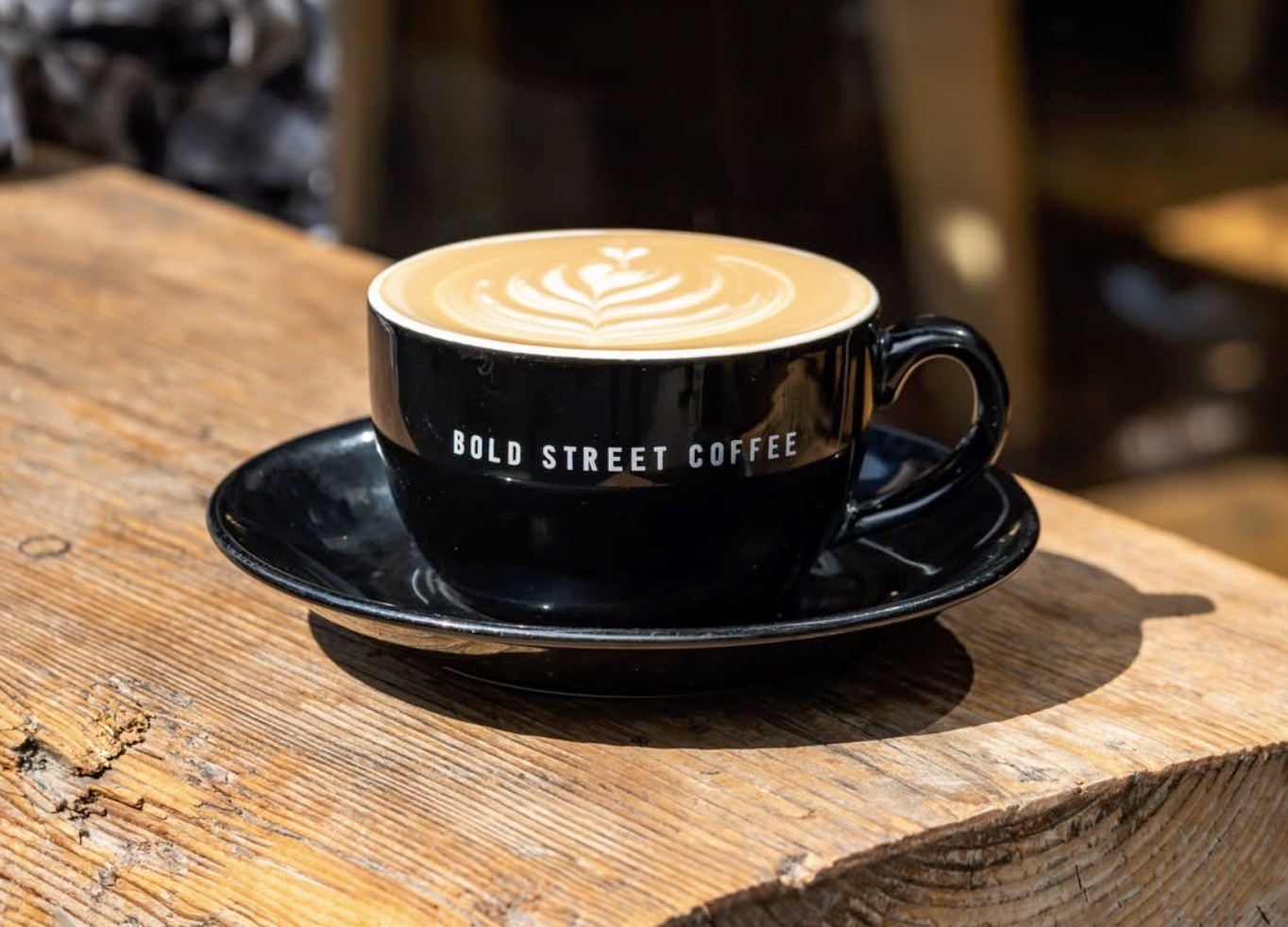 A black branded Bold Street Coffee cup and saucer with leaf latte art on a sunlit wooden table