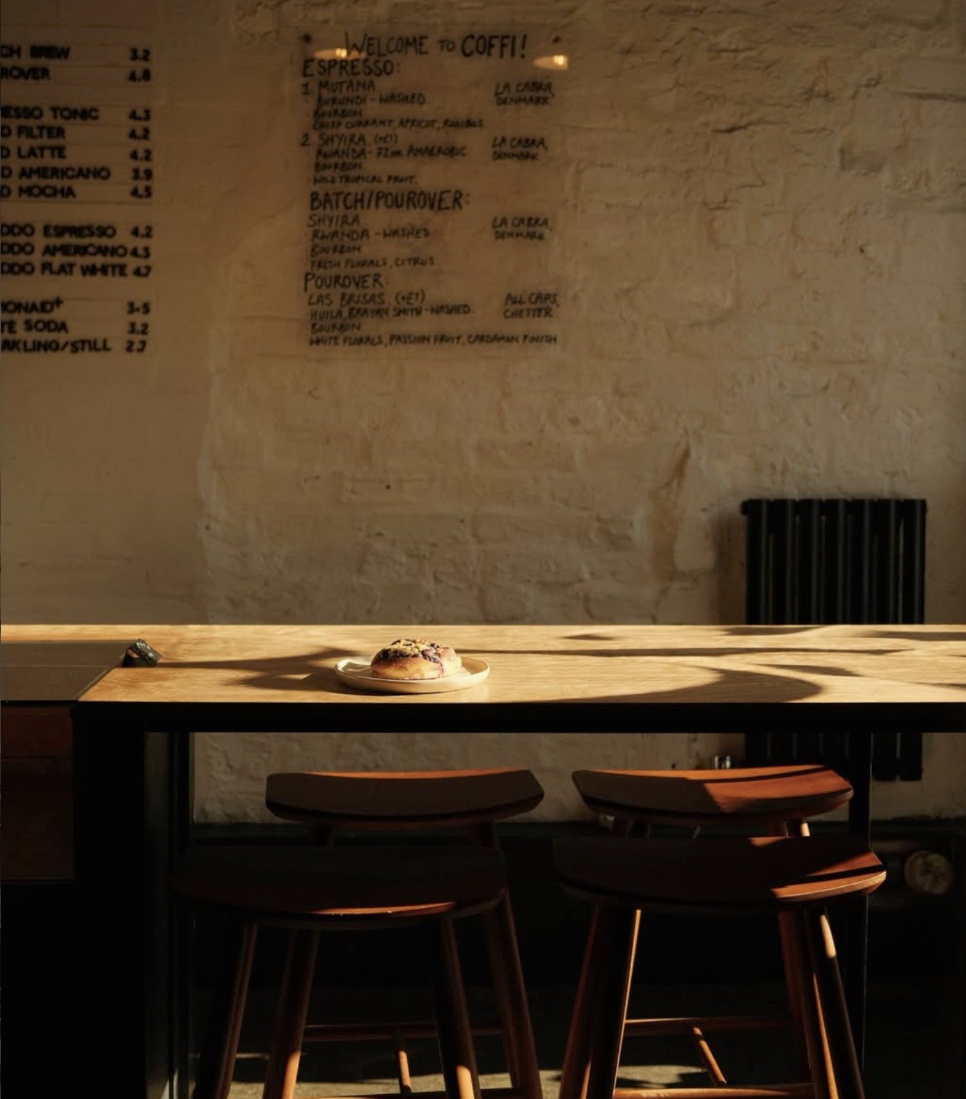 A handwritten espresso and pourover menu on a whitewashed brick wall above a sunlit wooden communal table at COFFI