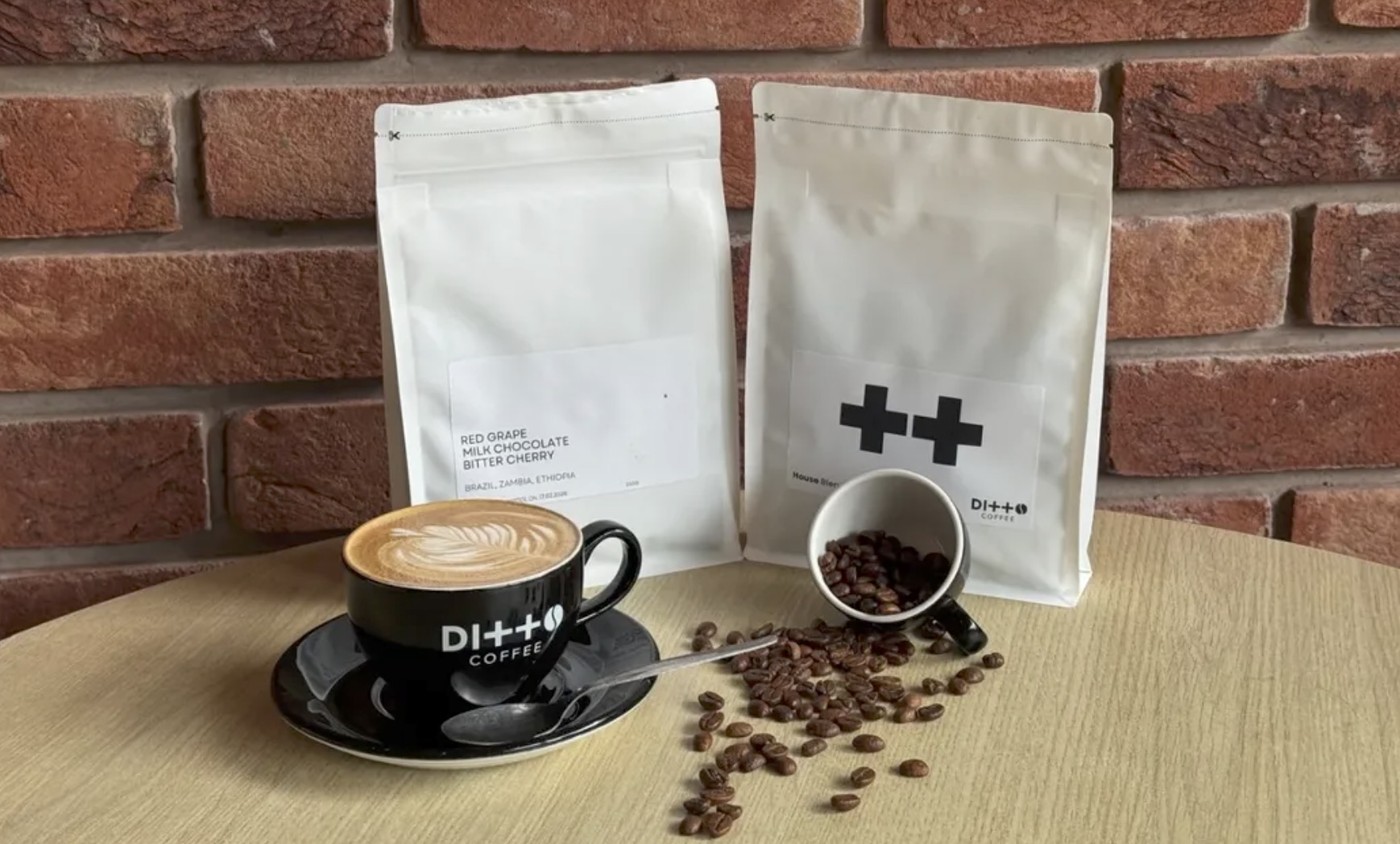 A branded Ditto Coffee cup with leaf latte art beside two retail bags and scattered coffee beans against a brick wall