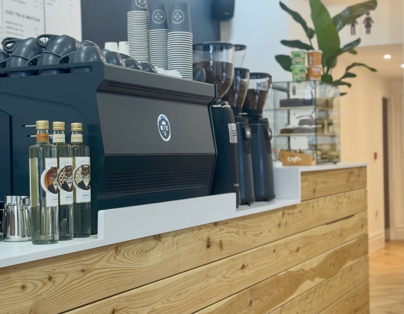 An espresso machine, grinders, and flavored syrups on a wood paneled counter at Rose Lane Coffee