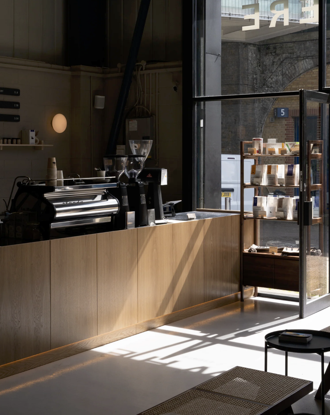The counter at Elsewhere's Deptford roastery bar, an espresso machine and retail bean bags on the shelf, tables lit by low sun