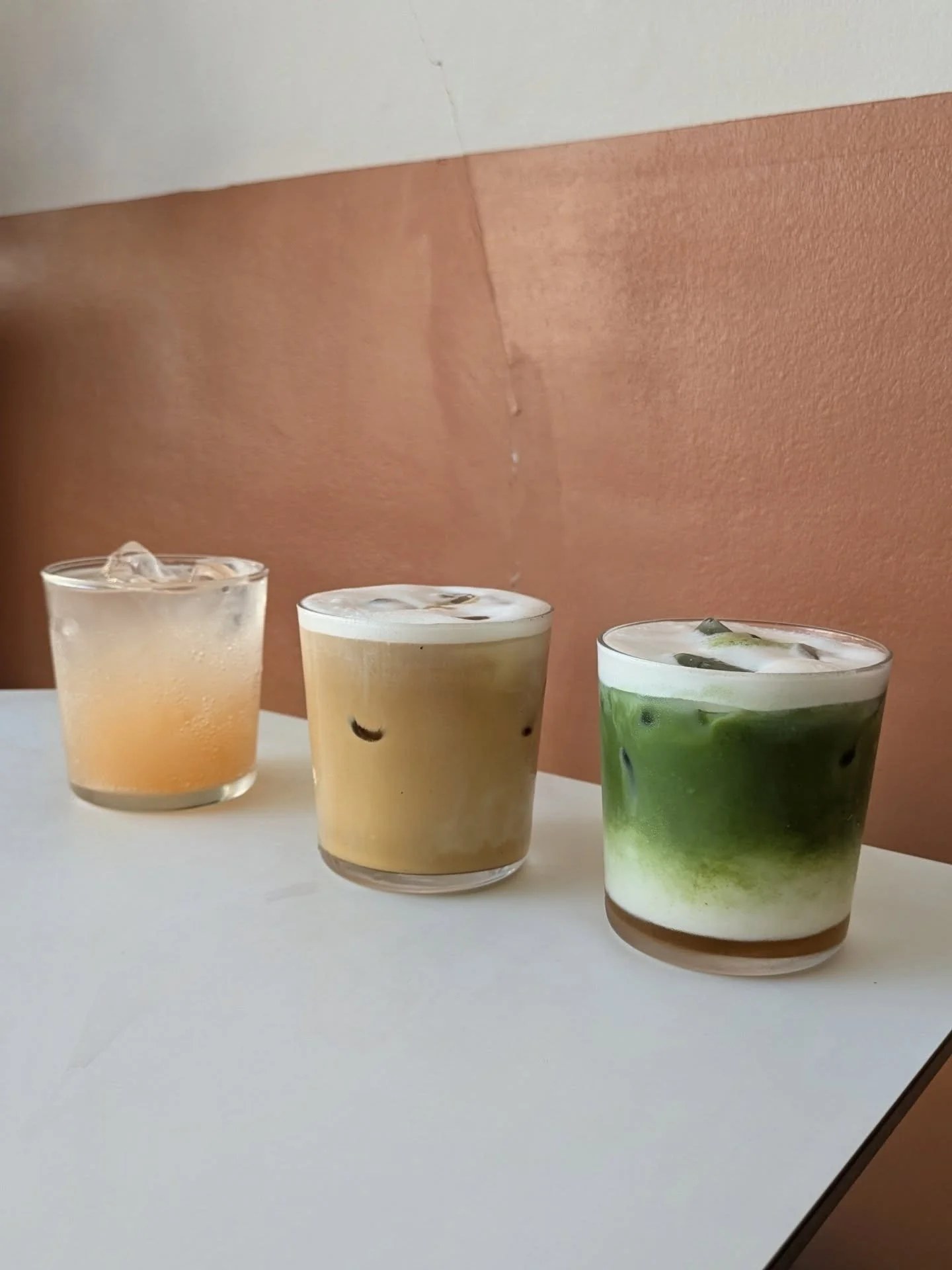 Three iced drinks on a table at Esters, an iced coffee and an iced matcha alongside a sparkling soda, against a terracotta wall