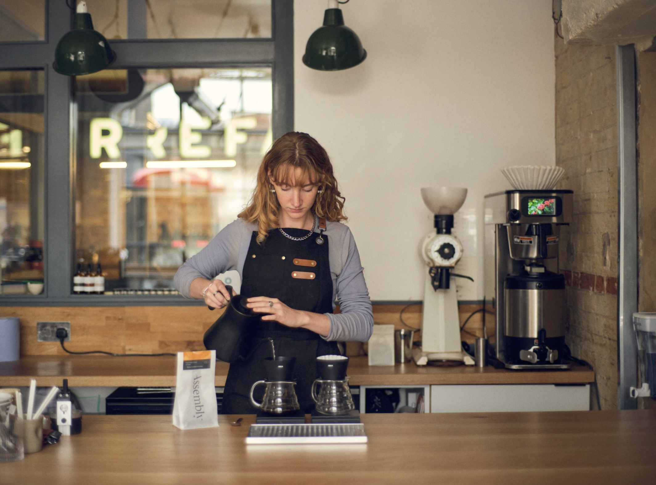 A barista pouring from a kettle over two pour over brewers on the counter at Door, roastery equipment behind