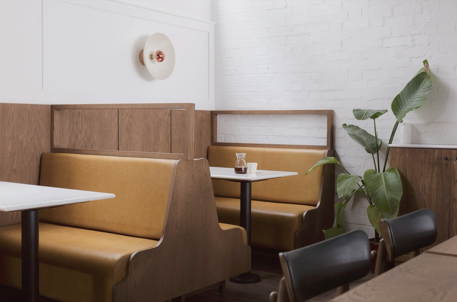 Mustard-leather booth seating at Prufrock Coffee, whitewashed brick, a plant, and a pour-over carafe on a marble table