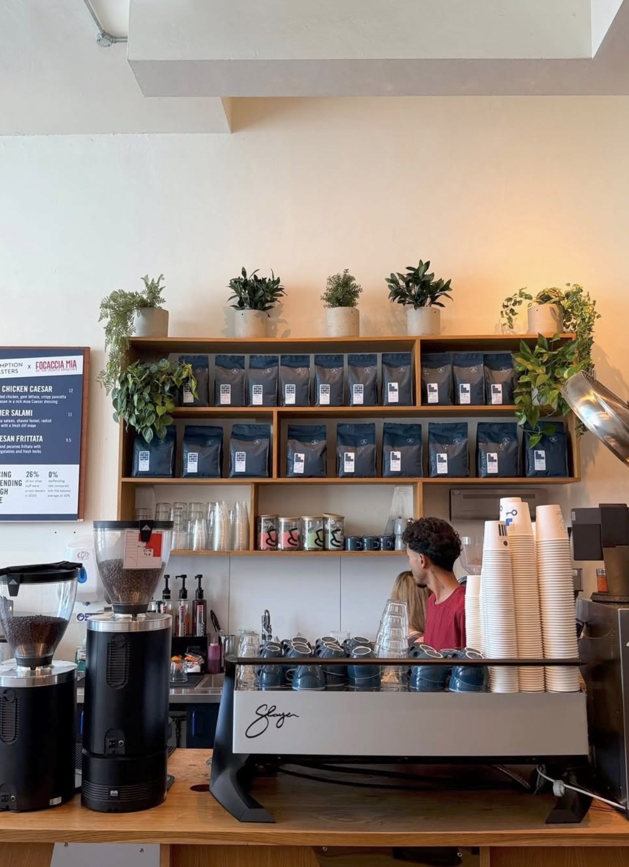 Shelves of retail coffee bags behind a Slayer espresso machine, a barista working with their back to the camera