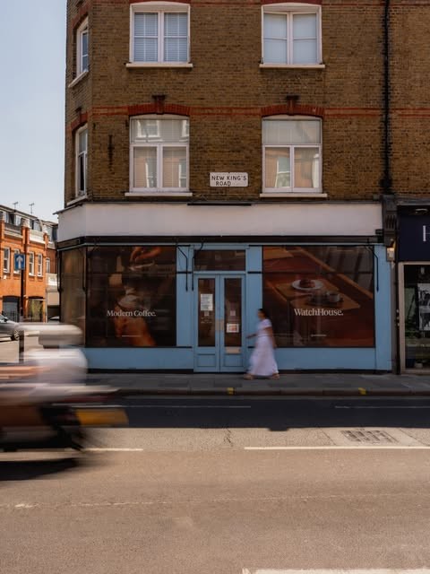 A blue fronted WatchHouse storefront with 'Modern Coffee. WatchHouse.' lettering on the window, a passerby blurred by motion on the pavement