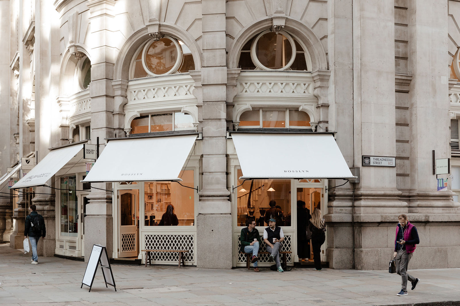 A white awninged Rosslyn storefront on a City of London corner, benches out front and the counter visible through the window