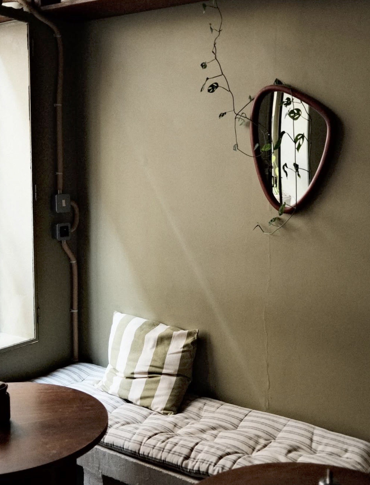 A striped cushion on a quilted window bench beneath a round mirror and trailing plant at MAGMA