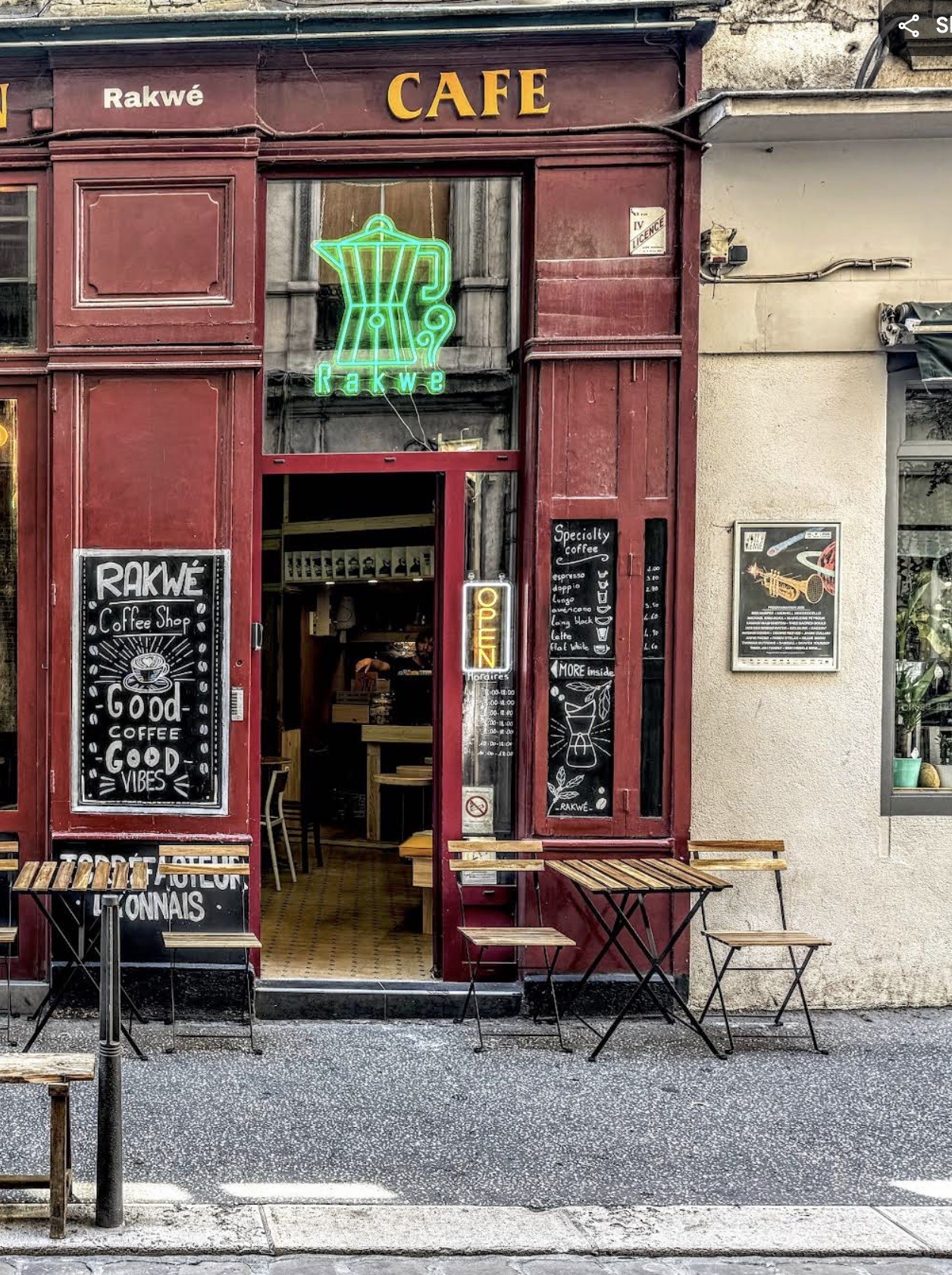 The red storefront of Rakwé with a glowing green neon moka pot sign and chalkboard menus beside outdoor bistro tables