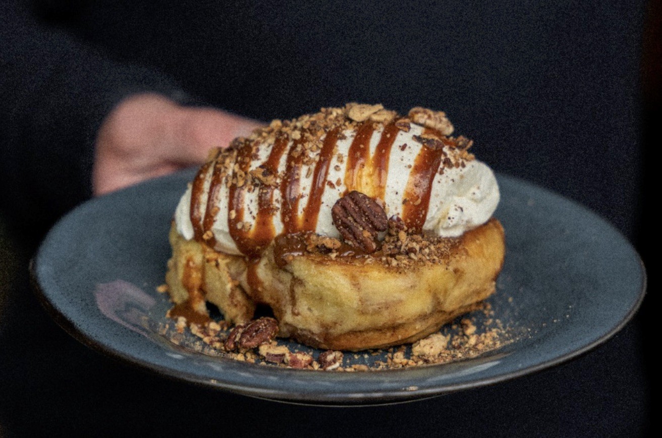 A hand presenting a plated brioche french toast topped with whipped cream, caramel, and pecans at Slake