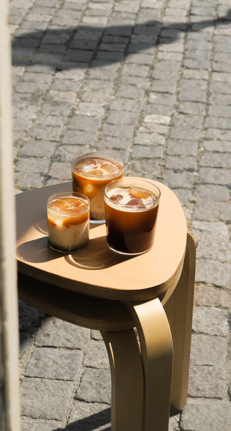 Three iced coffees in stubby glasses set on a wooden stool on the cobbled street outside