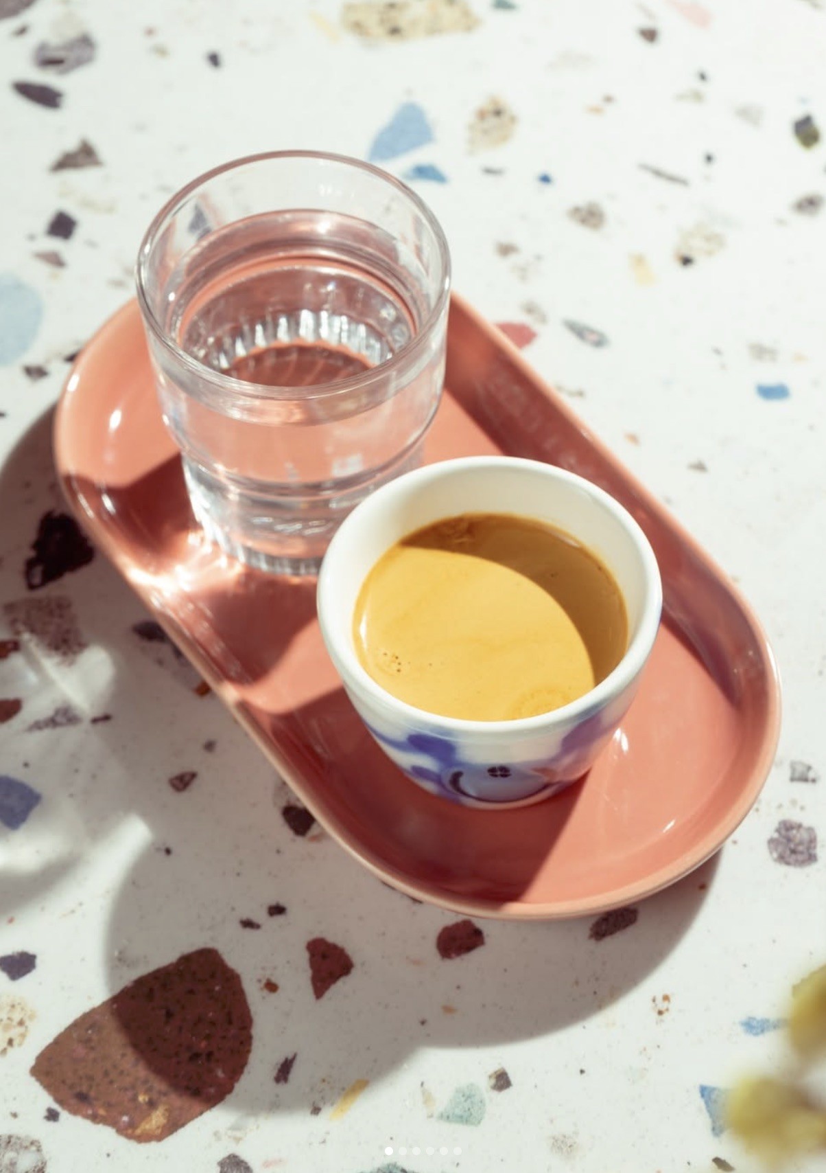 An espresso in a blue and white cup beside a glass of water on a pink tray, set on a speckled terrazzo table