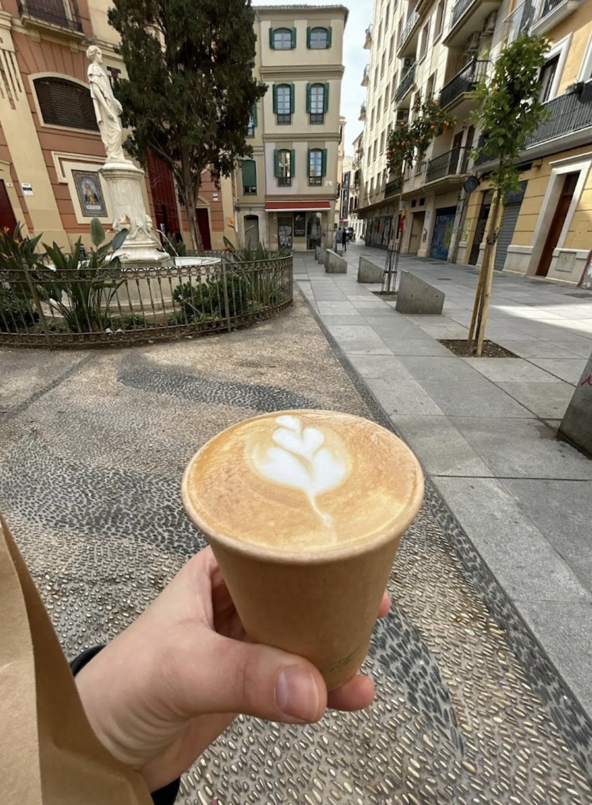 A takeaway cup of latte art held up against the view of Plaza de San Francisco outside the shop