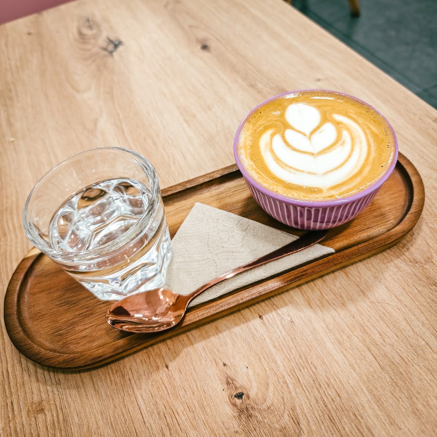 A latte with rosetta latte art in a striped purple bowl on a wooden tray, with a glass of water and a copper spoon