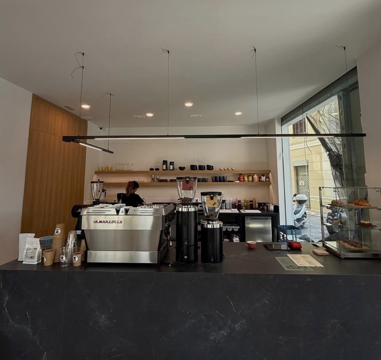 The espresso bar at Surge with a La Marzocco machine, grinders, and a pastry case beneath a wood-shelved back wall