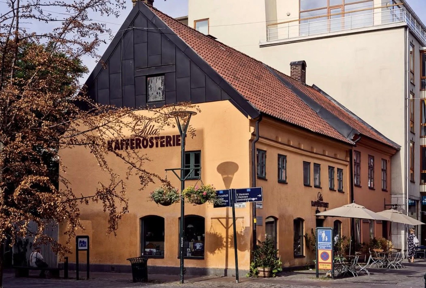 The honey colored corner building of Lilla Kafferosteriet with painted signage, hanging flower baskets, and outdoor umbrellas