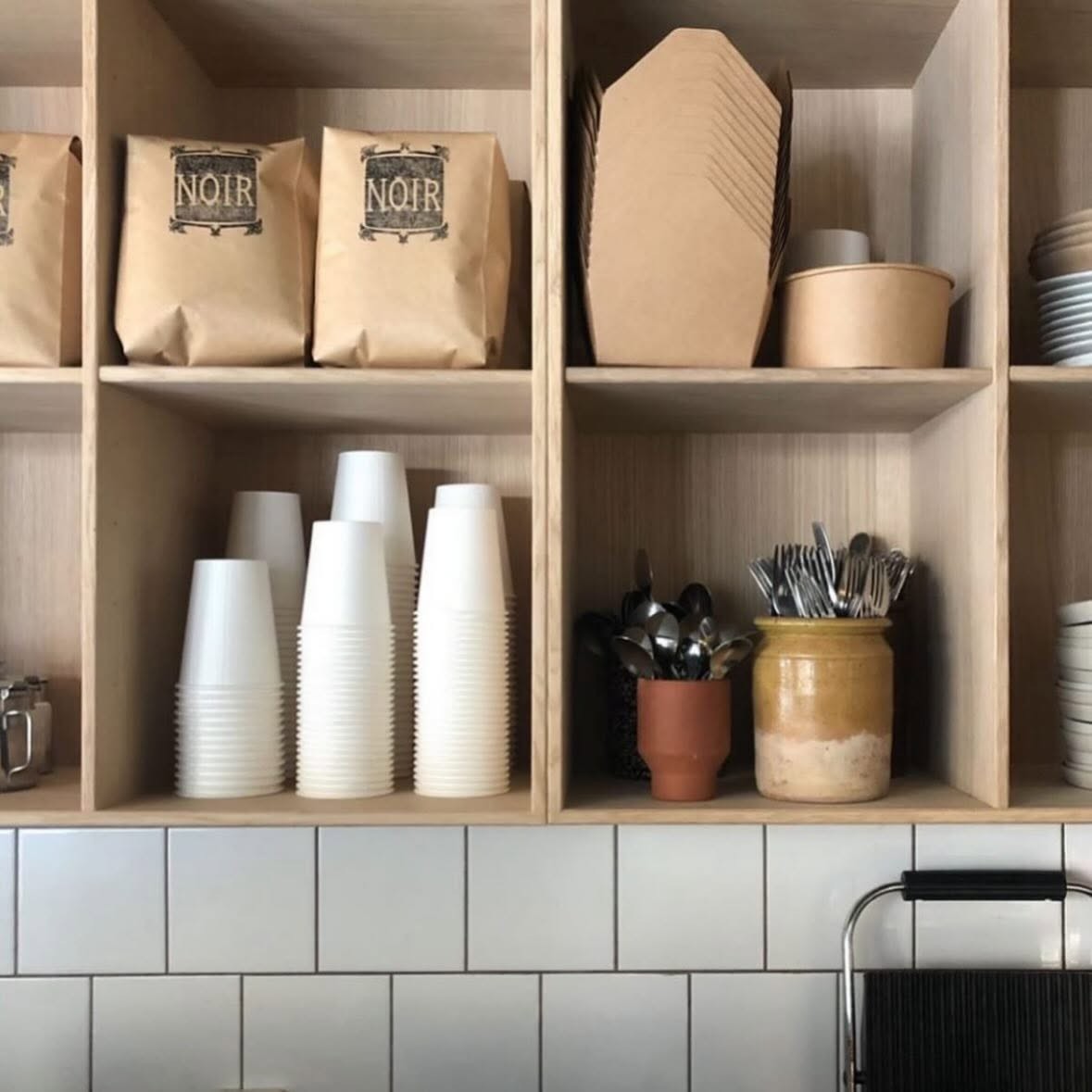 Retail bags of Noir house coffee beans on a shelf above stacked cups and jars of cutlery