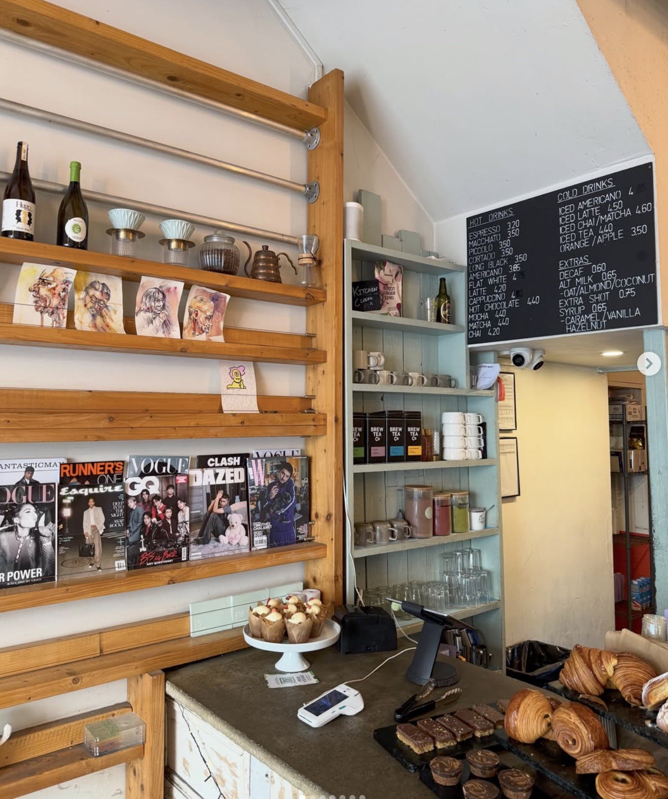 Wooden shelving stocked with magazines, artwork, and mugs beside the menu board and pastry counter at Fig + Sparrow