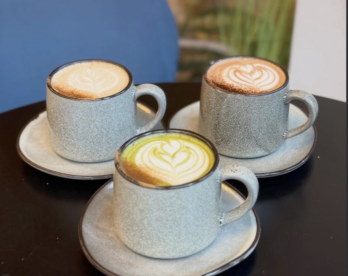 Three latte art drinks, including a matcha, served in speckled stoneware cups and saucers on a black table at Hampton & Vouis