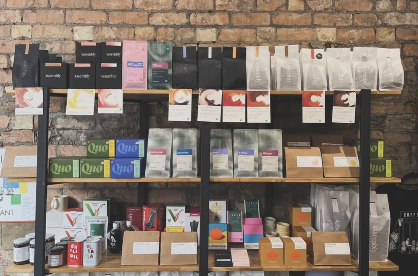 Retail shelves of coffee bags and brewing gear from Assembly, Quo, and other roasters against an exposed brick wall at Just Between Friends