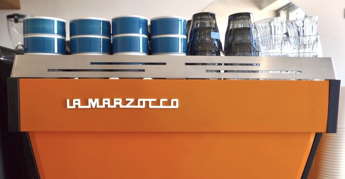 An orange La Marzocco espresso machine with rows of blue ceramic cups and glass tumblers arranged on top