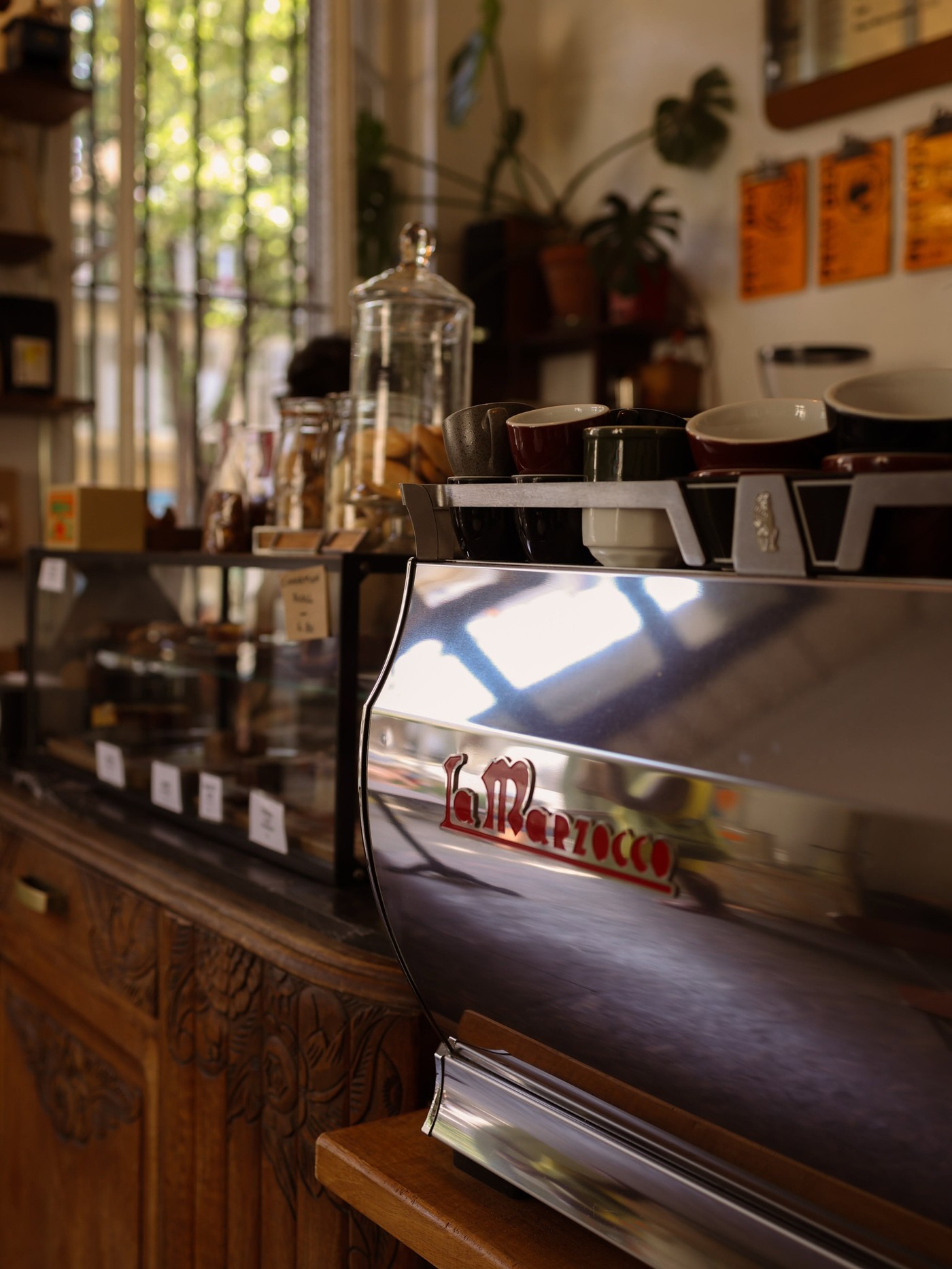 A La Marzocco espresso machine and grinder on a wooden bar top, with bags of house roasted beans and a display case of pastries on the shelf behind