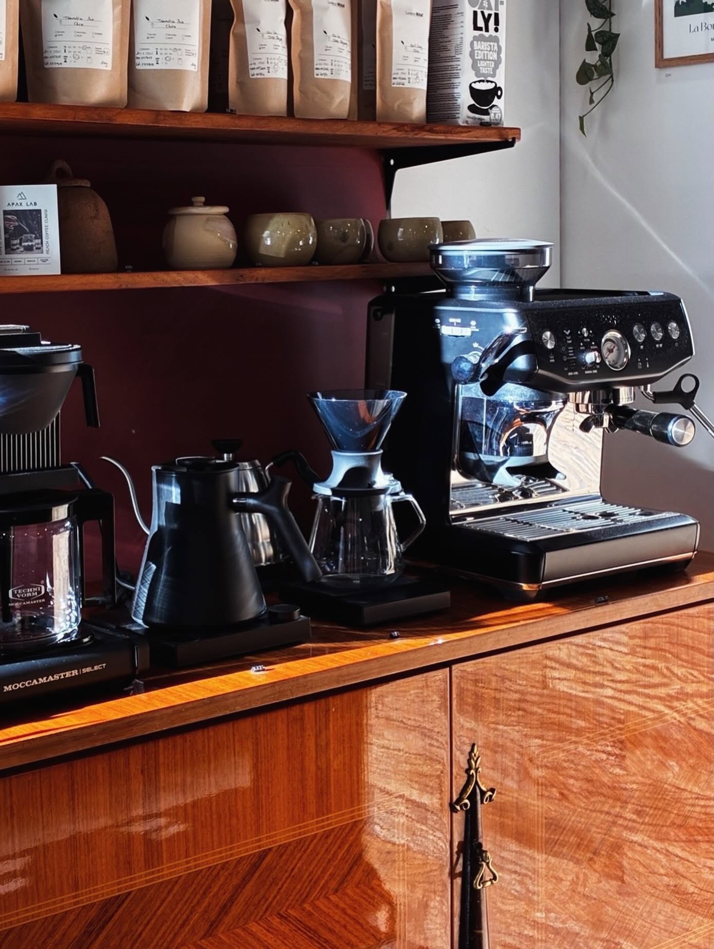 Bags of house roasted beans on a wooden shelf above a pour over bar with kettles, a dripper, and a black espresso machine and grinder