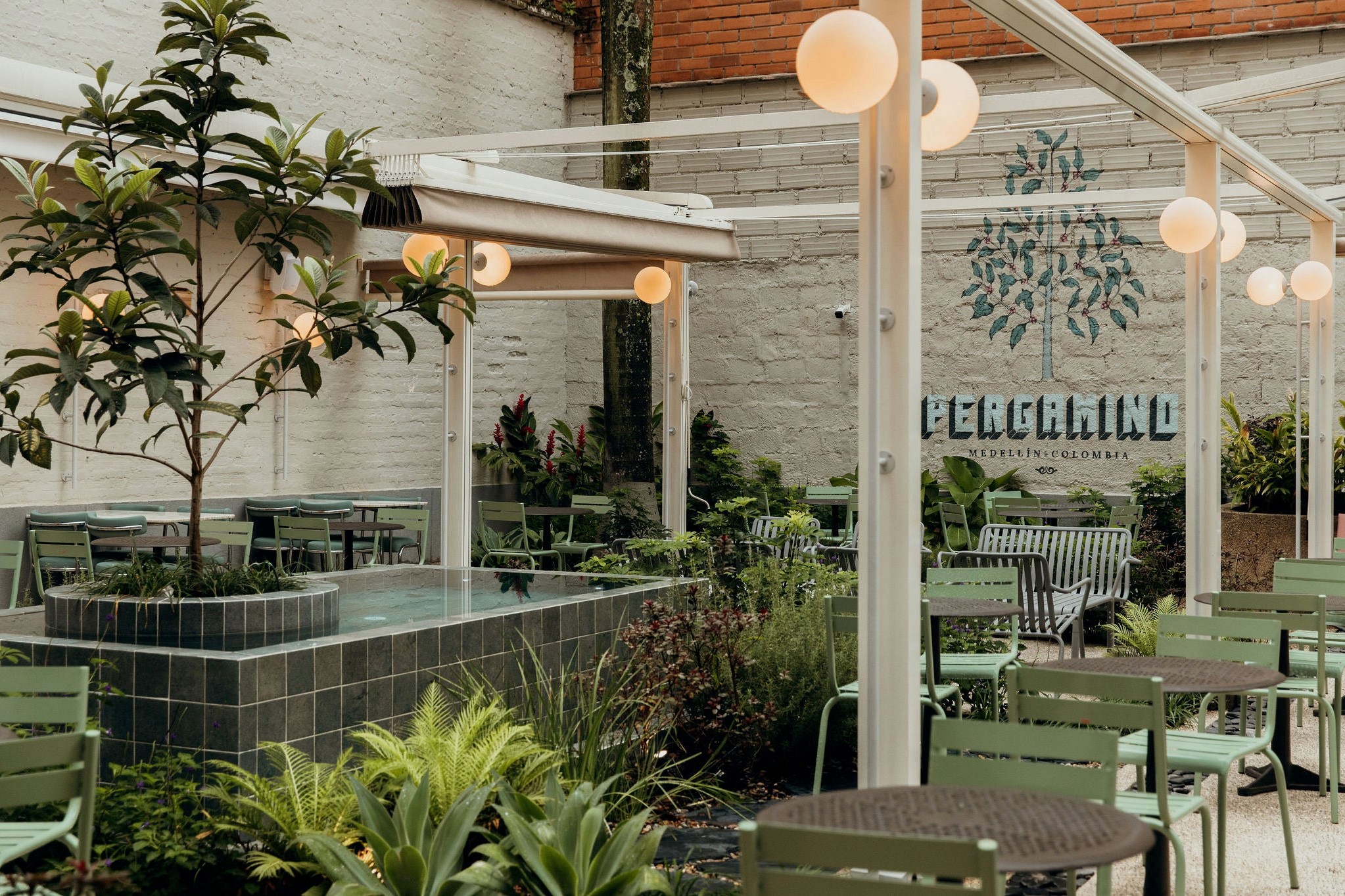 The courtyard garden at Pergamino Café Laureles, with the Pergamino coffee tree mural on a white brick wall, a tiled fountain and green café chairs