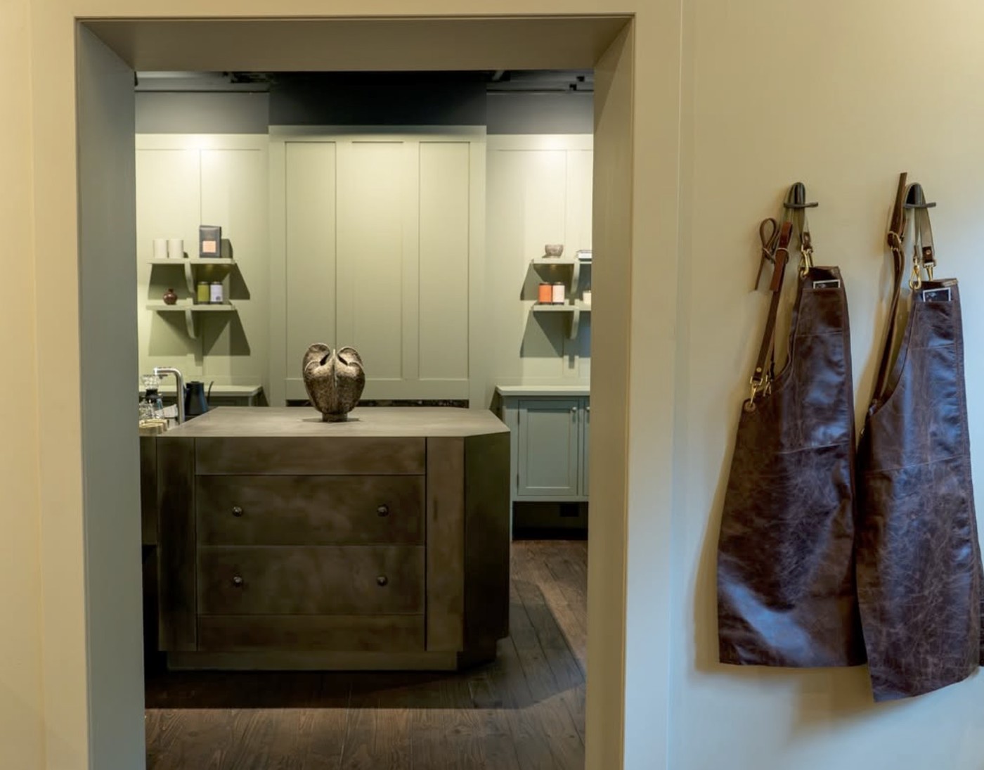 A private tasting room at Patricia with leather aprons hanging on the wall, sage green cabinetry, and a bronze sculpture on a dark island counter