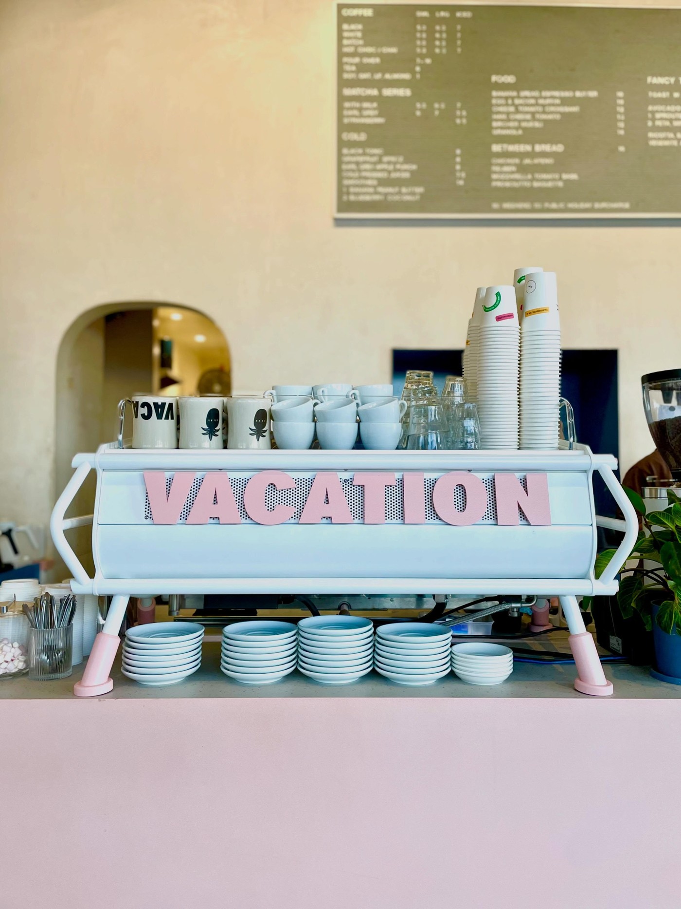 The pastel blue Vacation espresso machine with pink lettering, branded mugs, and stacked takeaway cups beneath the menu board