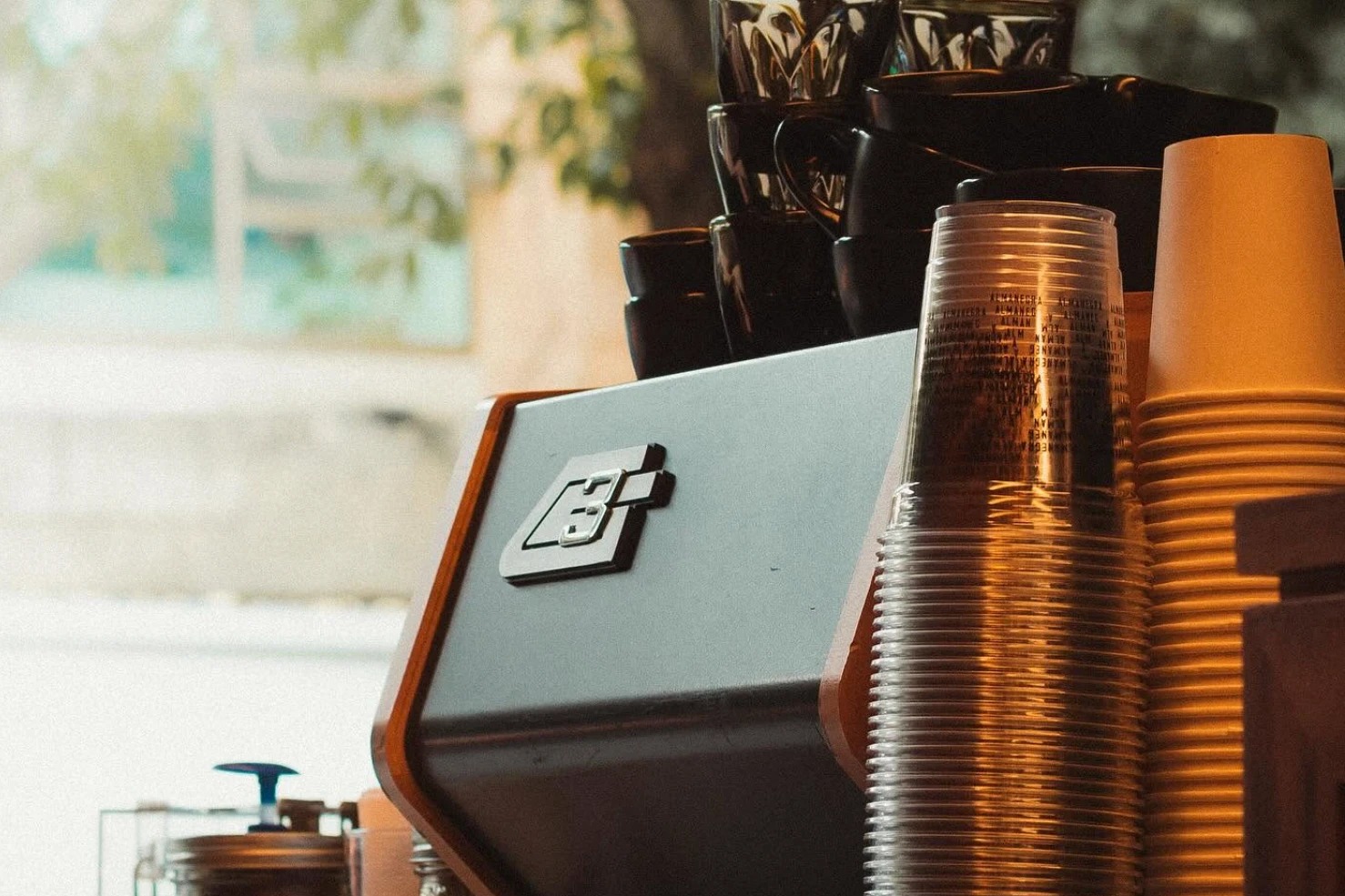 A bar machine with a sculpted copper trim and stacks of glass and black cups on top, beside towers of clear and paper takeaway cups in warm window light