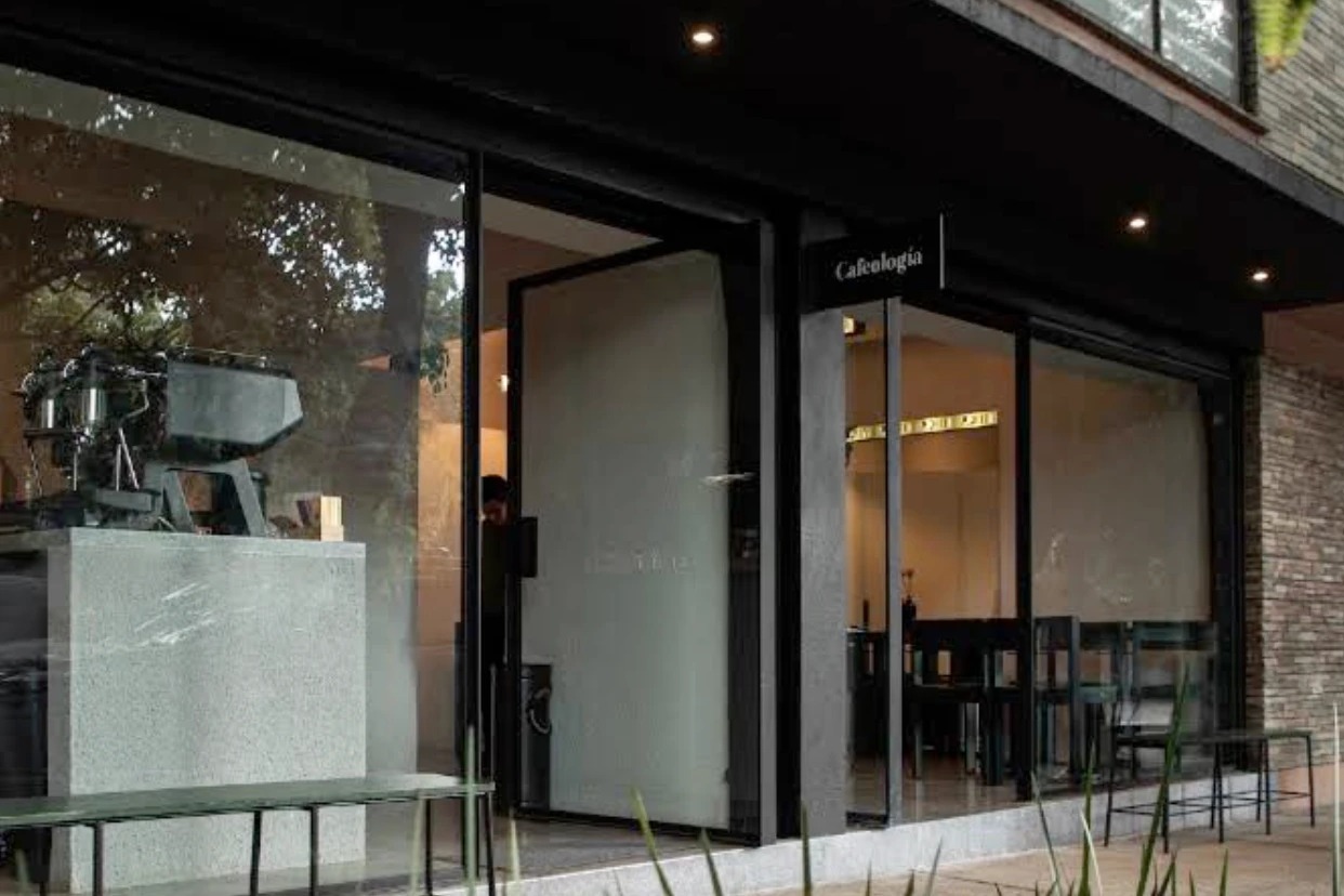 The glass fronted corner of Cafeologia Coyoacan under a dark overhang, the Cafeologia sign above the entrance, a pale terrazzo counter with an espresso machine inside and a bench outside