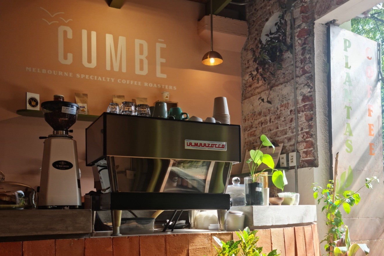 The espresso bar at Cumbé in Roma Norte, a La Marzocco machine and grinder under a CUMBÉ Melbourne Specialty Coffee Roasters wall sign, with a brick wall and plants beside a sunlit window