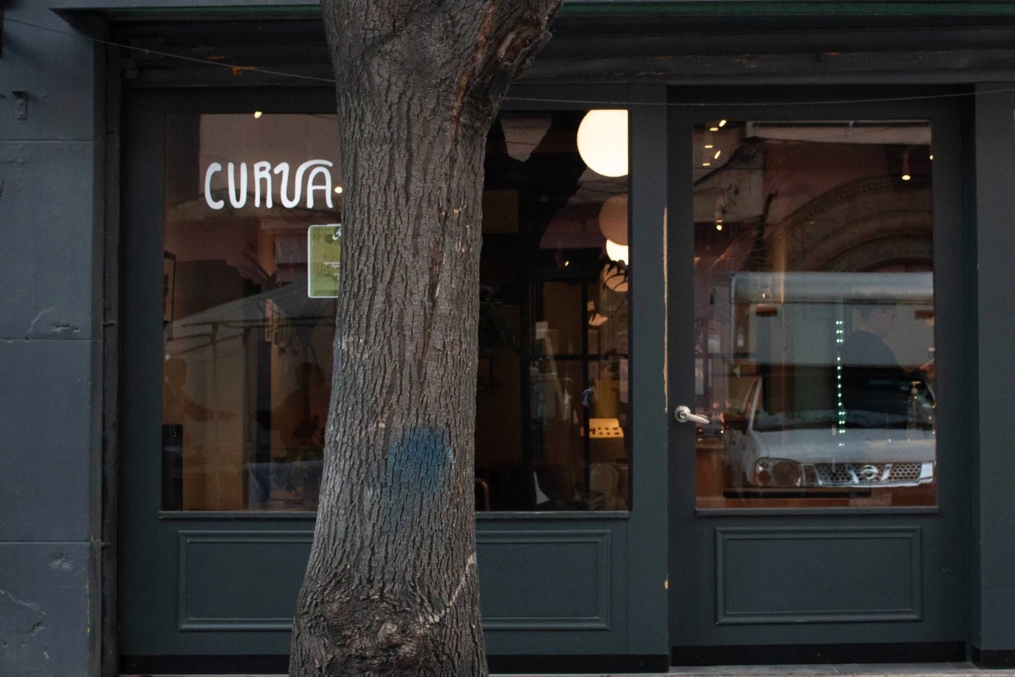 The dark green shopfront of Curva Cafe with the white Curva lettering on the window, round pendant lights glowing inside and a street tree in front