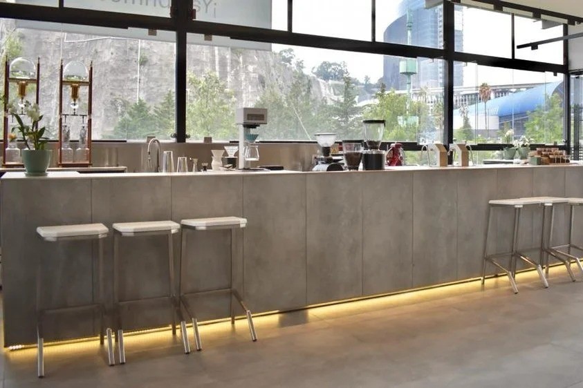 The long concrete finish coffee bar at Exploradores Club de Café in Santa Fe, with brewing equipment, a cold drip tower and bar stools in front of floor to ceiling windows