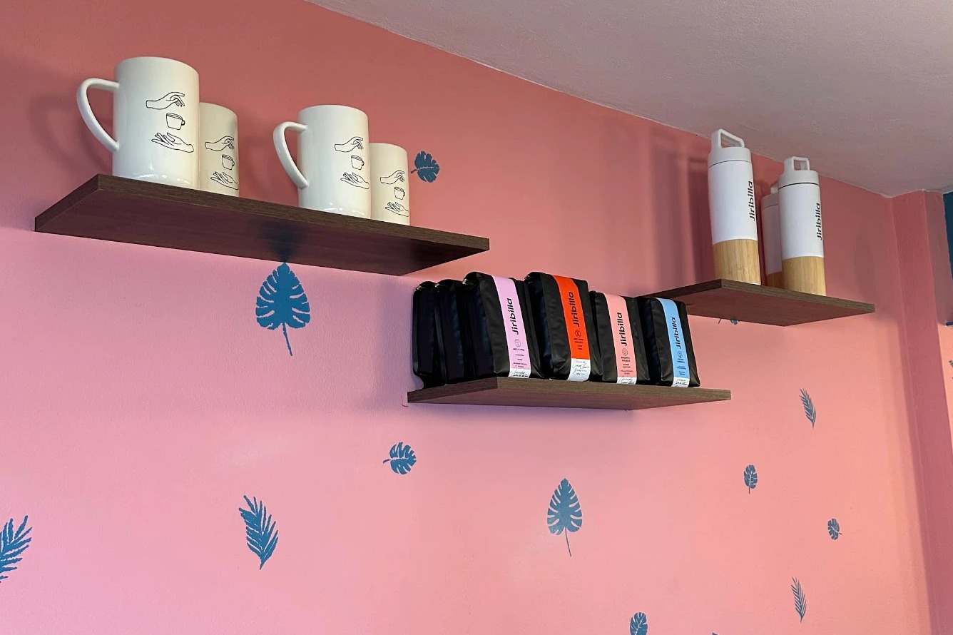 A pink wall with blue leaf prints and three dark wood shelves holding tall white mugs, white and wood flasks and rows of colourful Jiribilla coffee bags