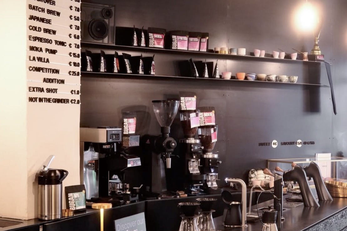 The Bugan Coffee Lab counter, a chalkboard style menu, grinders and pour over equipment beneath a shelf of retail bags and ceramic cups