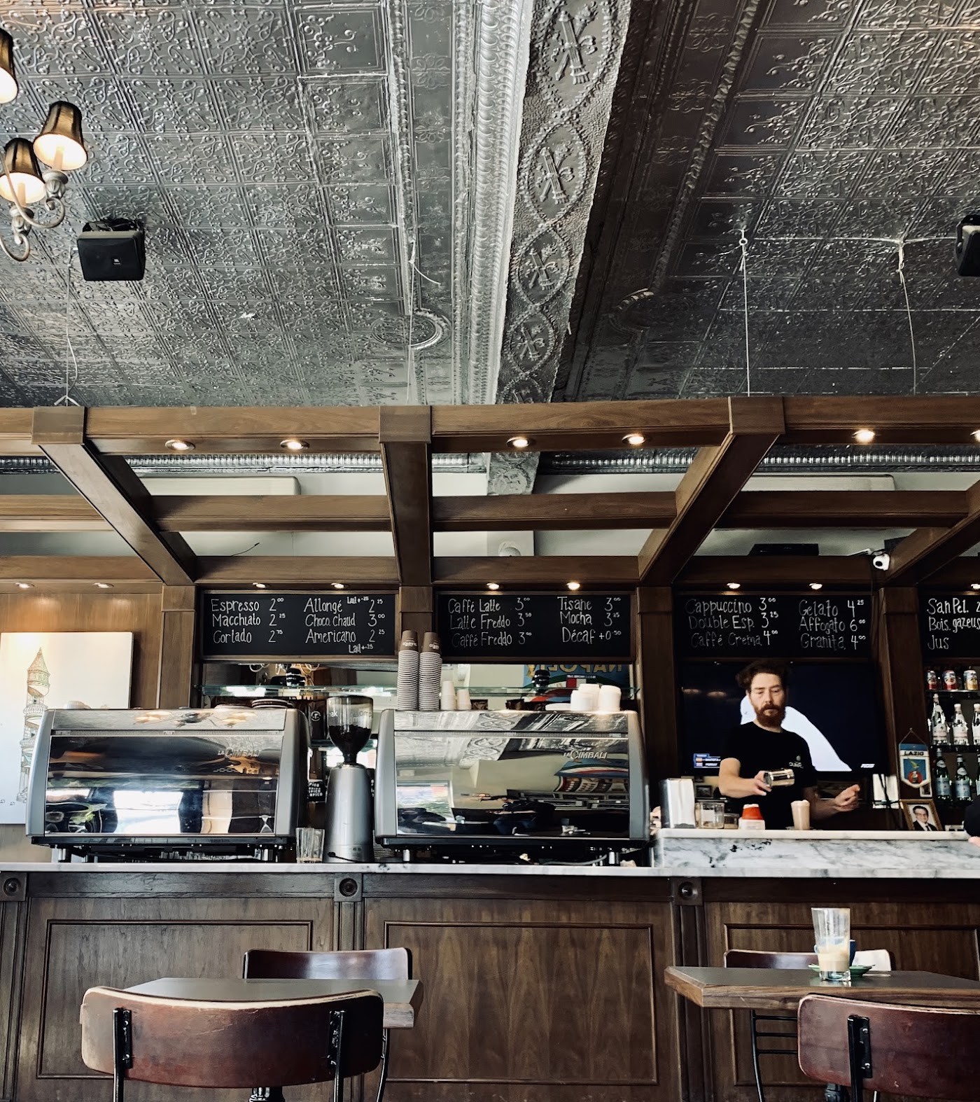 Behind the marble counter, a barista works between two La Cimbali espresso machines under an ornate pressed tin ceiling, with chalkboard menus listing espresso and cappuccino prices