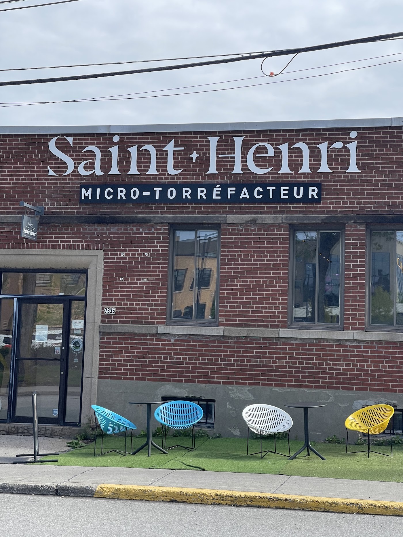 The brick facade of Café Saint-Henri's Mile-Ex headquarters, with the Saint-Henri Micro-Torréfacteur sign above the entrance and colorful patio chairs set out on artificial turf