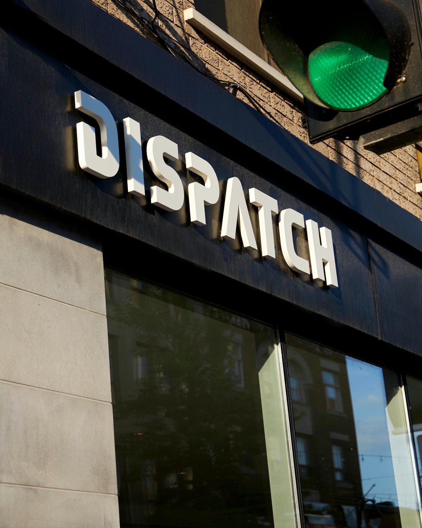 Close-up of the raised white Dispatch lettering on the storefront's dark awning, brick facade and a green traffic light visible above