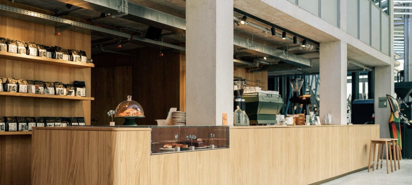 The wooden counter at Alrighty Coffee, retail bags on shelving and a green espresso machine behind the pastry case
