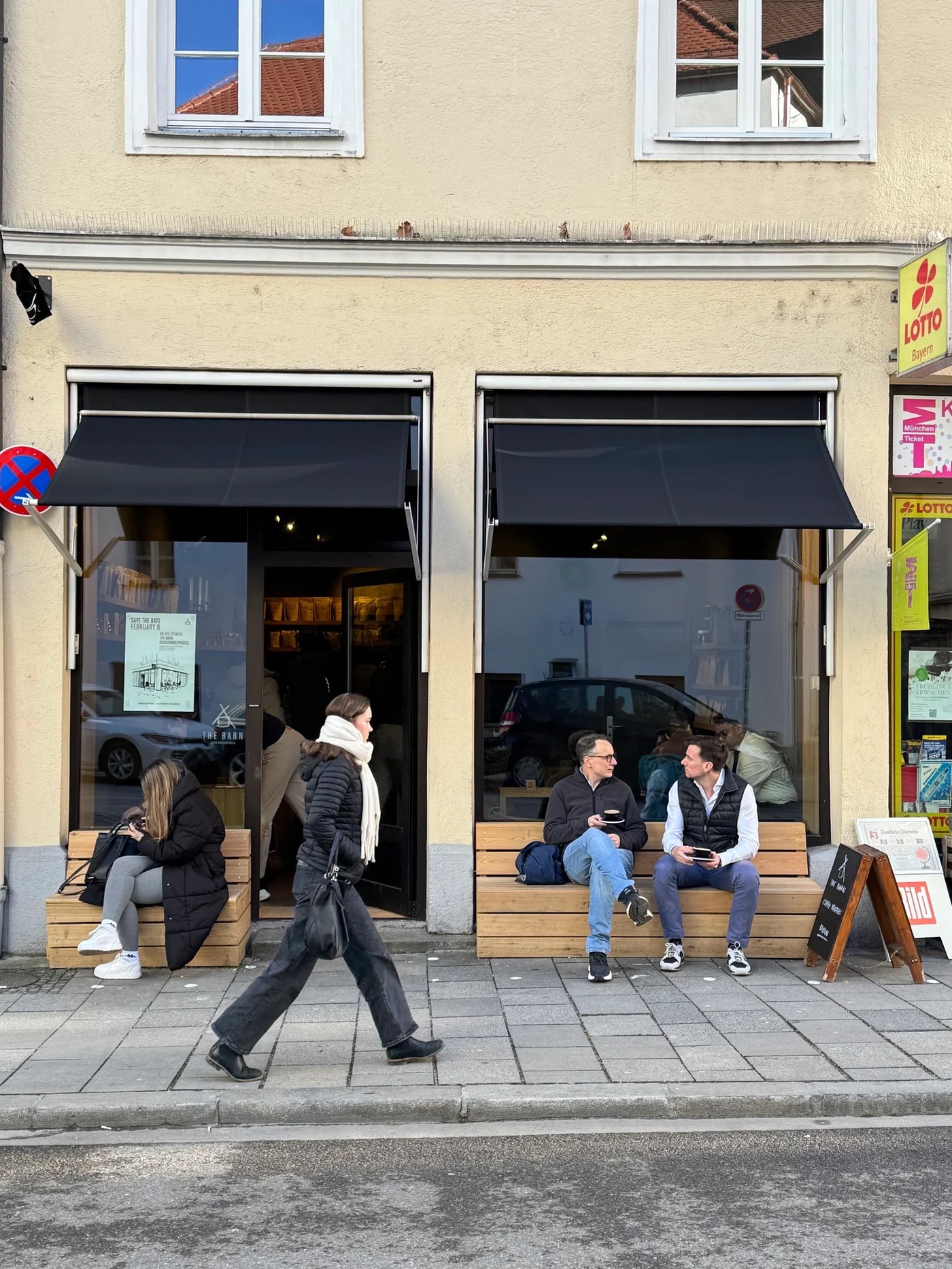 The Barn's Glockenbachviertel storefront with a hand drawn opening announcement poster in the window and customers seated on benches out front