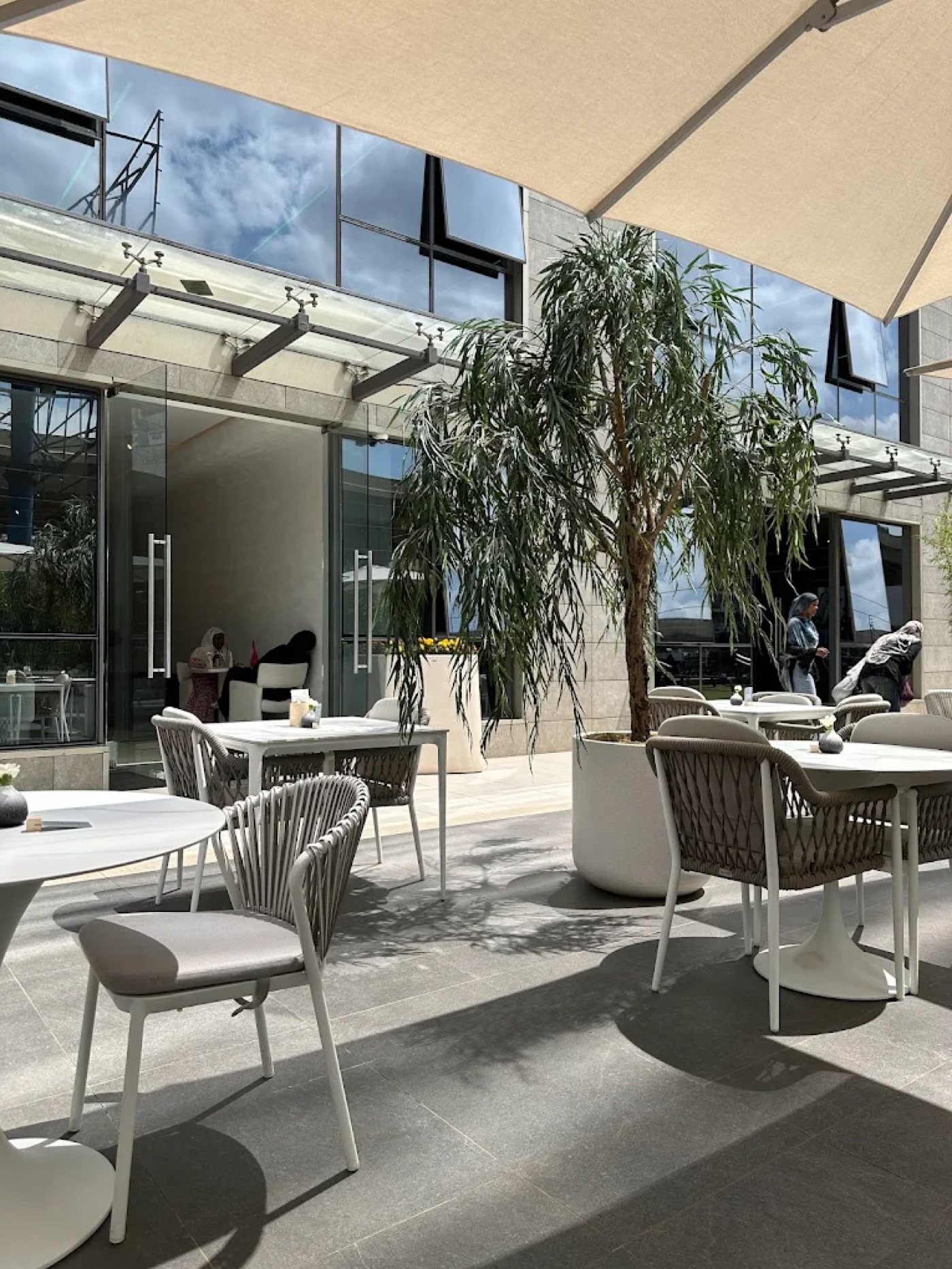 An outdoor patio at Ora Cafe with woven chairs, marble top tables, and a large potted tree under a canvas awning